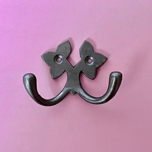 Puede incluir: Un gancho doble de metal negro con un diseño floral decorativo. El gancho está montado en una pared rosa.