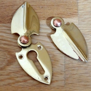 May include: A pair of brass door hinges with a copper accent. The hinges are shaped like a leaf and have a keyhole cutout.