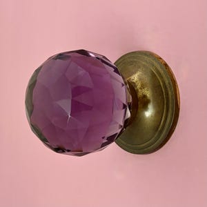 May include: A faceted, deep purple crystal doorknob with a brass base. The crystal is a rounded shape with many flat surfaces, reflecting light. The brass base is circular and has a slightly aged appearance. The background is a solid light pink.