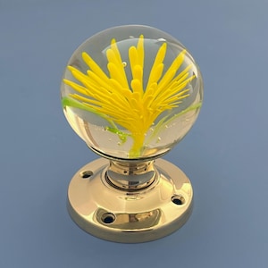 Op de afbeelding: Decoratieve deurknop met een heldere glazen bol met een geel bloemmotief en groene accenten. De basis is van gepolijst messing. Een uniek woondecoratie-item.