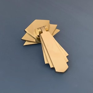 Peut inclure: Une poignée de porte en métal doré avec un design géométrique en forme d'éventail. La poignée a une poignée rectangulaire et une base décorative.
