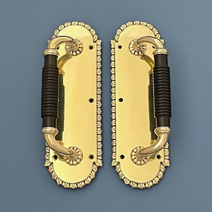 Puede incluir: Un par de manijas de puerta de latón dorado. Cada manija presenta una placa trasera ovalada con bordes decorativos y un agarre negro acanalado. Las manijas tienen un diseño curvo con detalles intrincados.