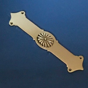 Puede incluir: Un tirador de puerta de latón con un diseño floral decorativo en el centro. El tirador es largo y rectangular con extremos redondeados.