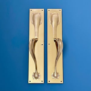 Puede incluir: Dos manijas de puerta de latón con un diseño curvo. Cada manija está montada en una placa rectangular con bordes redondeados y pequeños orificios para tornillos. Las manijas tienen un acabado pulido. El fondo es azul liso.