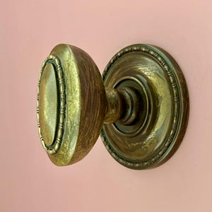 Könnte beinhalten: Ein runder Türknauf aus Messing mit verwittertem Antik-Finish und einer ovalen Mittelplatte. Der Knauf verfügt über einen dekorativen Perlenrand an der Kante und ist an einer passenden runden Rosette mit ähnlichem Perlenrand-Detail befestigt.