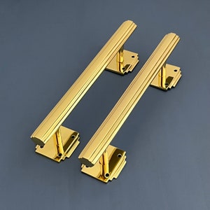 Può includere: Due maniglie per porte dorate con un design Art Deco. Le maniglie sono in ottone massiccio e hanno una finitura lucida.