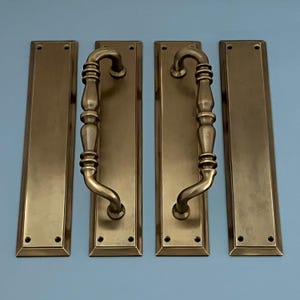 Puede incluir: Conjunto de tres tiradores de puerta de latón antiguo con un diseño decorativo. Cada tirador presenta una palanca larga y curva con un extremo redondeado y una sección decorativa acanalada en el medio. Los tiradores están montados en placas rectangulares con un borde ligeramente elevado.