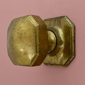 May include: A brass door knob and backplate, showing signs of age, set against a pink backdrop. The knob is octagonal, and the backplate is square with an octagonal frame. The brass has a worn, aged appearance.