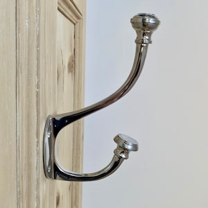 Puede incluir: Un gancho para abrigos de metal plateado con un diseño curvo y dos pomos redondeados en los extremos. El gancho está montado en una superficie de madera clara.