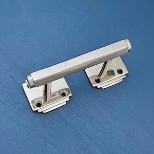 Op de afbeelding: Een gepolijste chromen deurgreep met een rechthoekige stang en twee vierkante platen. De platen hebben een geometrisch ontwerp met afgeronde hoeken.