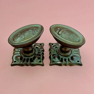 Könnte beinhalten: Ein Paar dunkel bronzefarbene Metall-Türknäufe mit ovalem Griff, der ein erhabenes Reliefdesign aufweist, und einer quadratischen Grundplatte mit vier Schraublöchern. Die Beschläge haben eine verwitterte Metalloberfläche mit sichtbaren dunkel- und hellgrünen Akzenten.