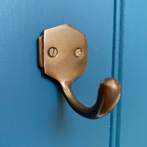 Op de afbeelding: Een bronzen metalen haak met een zeshoekige basis en een gebogen haak. De haak is met twee schroeven aan een blauwe muur bevestigd.