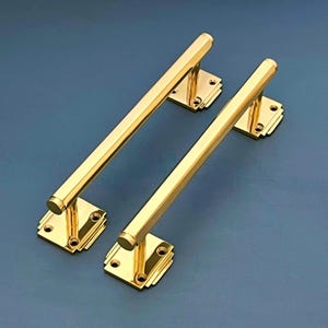 Può includere: Due maniglie per porte dorate con una base rettangolare e una lunga barra dritta. Le maniglie hanno un design minimalista art déco.