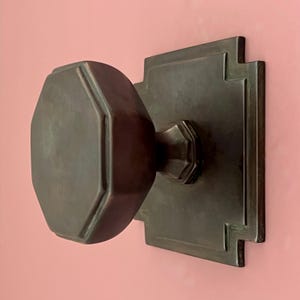 Puede incluir: Pomo de puerta en tono bronce con manija octogonal y placa trasera rectangular. La placa trasera tiene un diseño escalonado decorativo. El pomo está montado en una pared rosa, creando un contraste de color y textura.
