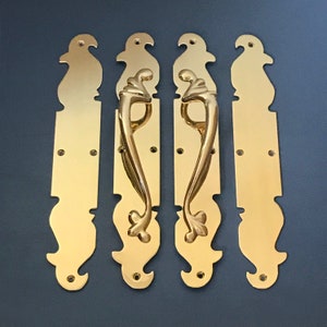 Pode incluir: Quatro puxadores de porta em latão polido. Duas pegas têm um design curvo com um elemento floral decorativo. As outras duas são placas traseiras retangulares com uma parte superior e inferior decorativas. As pegas e as placas traseiras são de cor dourada brilhante.