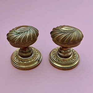 May include: A pair of oval brass door knobs featuring a swirling ribbed pattern on the top surface and a beaded border. Each knob is attached to a circular base plate with a matching tiered design and a decorative rope-like edge detail.