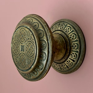 May include: Close-up of an antique brass doorknob featuring detailed floral and scrollwork. The doorknob has a circular design with a raised centre and a warm, aged patina. The background is a soft pink.