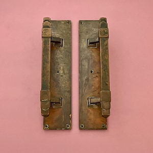 May include: Two antique brass door handles with a geometric design. The handles are rectangular with a raised, textured design and have a patina finish.