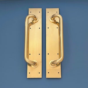 May include: Two gold door handles with a curved design. The handles are mounted on rectangular plates.
