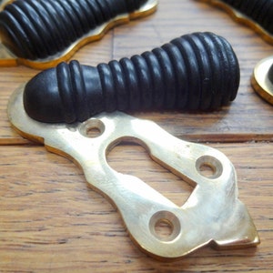 Op de afbeelding: Antieke zwarte en messing hardware set. De set bevat een zwarte, geribbelde handgreep en een messing sleutelgatplaat. De sleutelgatplaat heeft een decoratieve vorm met drie gaten. De hardware is waarschijnlijk voor een kast of lade.