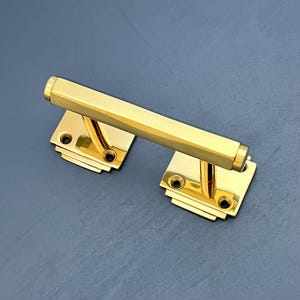 Può includere: Una maniglia per porta in oro lucido con una barra rettangolare e due piastre quadrate. La maniglia ha un design minimalista con bordi arrotondati.