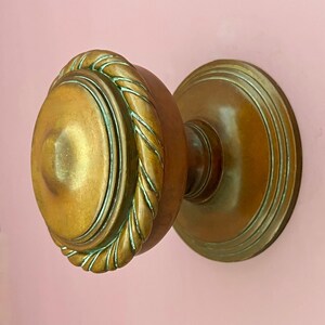 Könnte beinhalten: Ein runder Türknauf aus Messing mit einem markanten, seilartigen gedrehten Rand und einer glatten, leicht gewölbten Mitte. Das Beschlagteil hat ein warmes, gealtertes Metallfinish mit subtilen grünen Patina-Akzenten in den Vertiefungen.