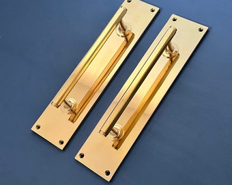 Könnte beinhalten: Zwei goldfarbene Metall-Türgriffe mit rechteckiger Form und einem langen, geraden Balken. Die Griffe sind auf einer ebenen Fläche montiert.