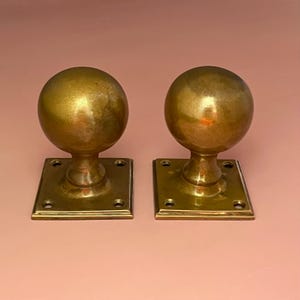 Puede incluir: Dos pomos de puerta antiguos de latón. Cada pomo tiene forma esférica sobre una base cuadrada. El latón tiene una pátina cálida y envejecida. Los pomos se muestran sobre un fondo rosa suave.