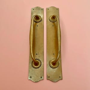 Puede incluir: Dos manijas de puerta antiguas de latón. Cada manija está montada en una placa rectangular con bordes redondeados y orificios para tornillos. Las manijas tienen un diseño curvo y un tono dorado cálido, mostrando signos de edad.