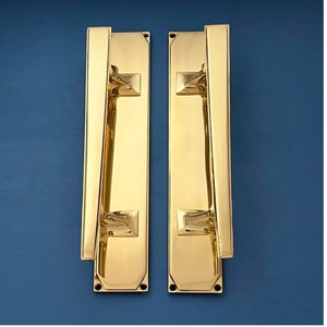 Puede incluir: Dos tiradores de puerta de latón pulido. Cada tirador tiene una placa trasera rectangular con un asa vertical y detalles cuadrados. El latón tiene un acabado brillante y reflectante. El fondo es azul oscuro.