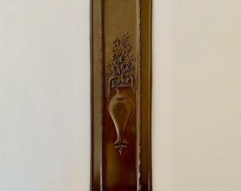 May include: A brown metal wall plaque featuring a raised design of a vase with flowers. The plaque is rectangular with a decorative border.