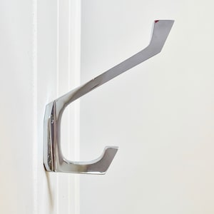 May include: A chrome-plated metal hook with a double-prong design. The hook is mounted on a white wall.