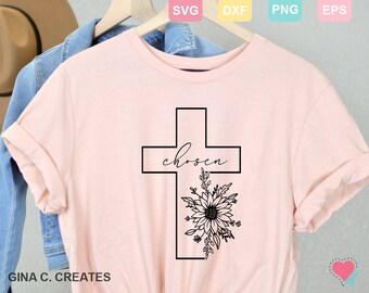 Può includere: Una maglietta rosa con un contorno nero di una croce e la parola "chosen" all'interno della croce. Un disegno floreale nero si trova sotto la croce.