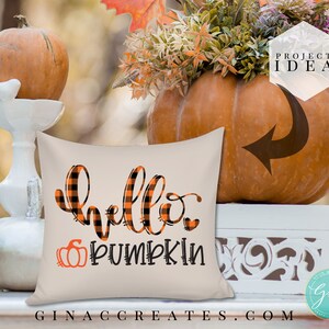 Op de afbeelding: Een beige kussen met de tekst "hello pumpkin" in zwart met een zwart-oranje buffalo plaid patroon. Het kussen ligt op een wit oppervlak met een pompoen en herfstdecoratie.