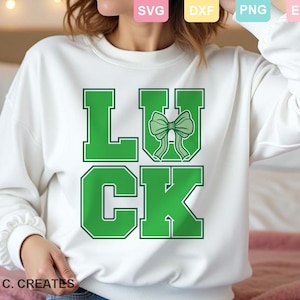 Pode incluir: Camisola branca com a palavra "LUCK" em letras verdes. O "U" apresenta um laço verde. Um estilo casual, perfeito para o Dia de São Patrício ou para o dia a dia. O texto "GINA C. CREATES" está na parte inferior.