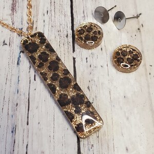 Op de afbeelding: Gouden ketting met een lange rechthoekige hanger en bijpassende oorbelletjes. De hanger en oorbelletjes hebben een bruin en gouden luipaardprint met glitter.
