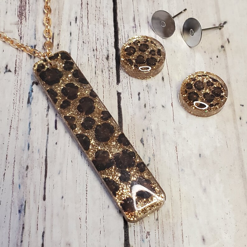 Op de afbeelding: Gouden ketting met een lange rechthoekige hanger en bijpassende oorbelletjes. De hanger en oorbelletjes hebben een bruin en gouden luipaardprint met glitter.