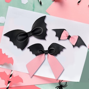 May include: Three decorative hair bows shaped like bat wings. One large solid black bow, one small bow with black wings and a light pink center, and one medium bow with black wings and two long light pink ribbon tails extending downward.
