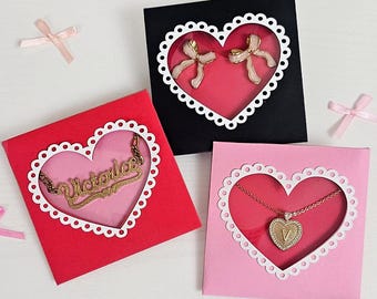 Può includere: Tre scatole regalo per San Valentino con ritagli a forma di cuore. Le scatole sono rossa, nera e rosa. La scatola rossa contiene una collana d'oro con il nome "Victoria" all'interno. La scatola nera contiene un paio di orecchini a fiocco rosa all'interno. La scatola rosa contiene una collana d'oro con un ciondolo a forma di cuore all'interno.