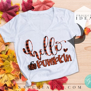 Op de afbeelding: Wit T-shirt met een zwart-oranje geruit ontwerp met de tekst "hello pumpkin" en een kleine pompoenafbeelding. Het shirt heeft een V-hals en een ontspannen pasvorm.