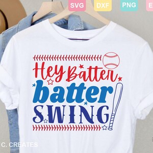 May include: White t-shirt with the text "Hey Batter Batter Swing" in red and blue, with a baseball and bat graphic. The design includes baseball stitching and stars. The shirt is hanging on a wooden hanger.