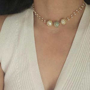 Puede incluir: Un delicado collar gargantilla con una doble hilera de peque&ntilde;as perlas blancas. El collar est&aacute; adornado con tres cuentas decorativas m&aacute;s grandes: dos con superficies de perlas y una con un dise&ntilde;o turquesa y dorado. El collar es dorado.