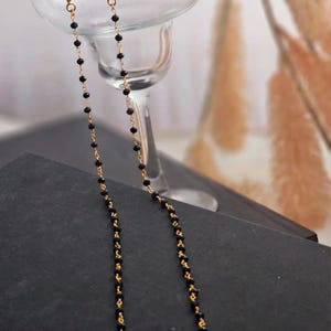 Può includere: Orecchini color oro con un lungo design pendente. Ogni orecchino presenta una serie di piccole perle nere infilate lungo una delicata catena. Gli orecchini sono esposti davanti a un bicchiere da margarita, con uno sfondo scuro.