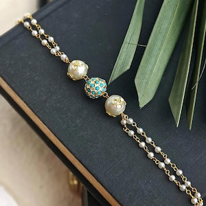 Puede incluir: Una pulsera dorada con tres cuentas decorativas. La cuenta central es turquesa, rodeada de cuentas m&aacute;s peque&ntilde;as. Las otras dos cuentas son blancas con detalles transparentes. La pulsera est&aacute; sobre un fondo oscuro.