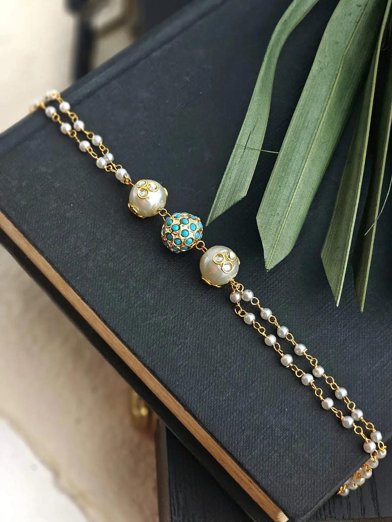 Puede incluir: Una pulsera dorada con tres cuentas decorativas. La cuenta central es turquesa, rodeada de cuentas m&aacute;s peque&ntilde;as. Las otras dos cuentas son blancas con detalles transparentes. La pulsera est&aacute; sobre un fondo oscuro.