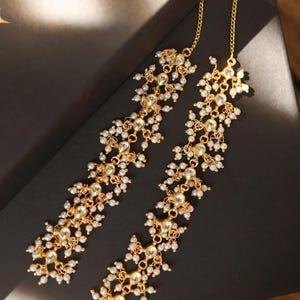 May include: Gold-toned earrings with a floral design, featuring clear gemstones and small white pearls. The earrings are displayed on a dark surface, showcasing their intricate details and elegant style.