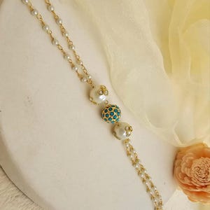 Puede incluir: Un collar dorado con una cadena delicada y peque&ntilde;as perlas blancas. El collar presenta tres cuentas decorativas m&aacute;s grandes: dos cuentas blancas con forma de perla con detalles dorados y una cuenta central de color turquesa. El fondo es blanco roto.