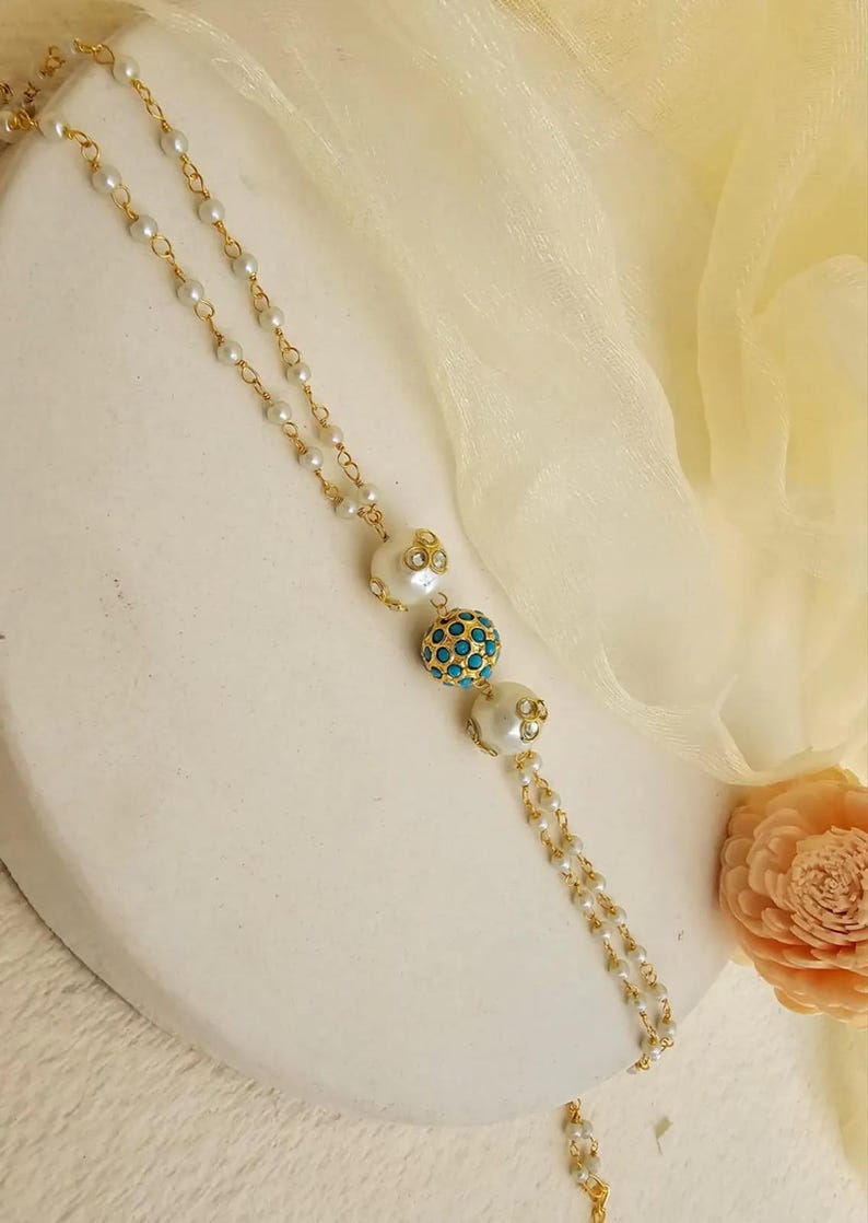 Puede incluir: Un collar dorado con una cadena delicada y peque&ntilde;as perlas blancas. El collar presenta tres cuentas decorativas m&aacute;s grandes: dos cuentas blancas con forma de perla con detalles dorados y una cuenta central de color turquesa. El fondo es blanco roto.