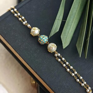 Puede incluir: Una pulsera dorada con tres cuentas decorativas. La cuenta central es turquesa, rodeada de cuentas m&aacute;s peque&ntilde;as. Las otras dos cuentas son blancas con detalles transparentes. La pulsera est&aacute; compuesta por peque&ntilde;as perlas blancas ensartadas en una cadena dorada.