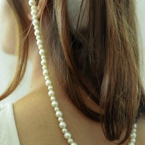 May include: A strand of white faux pearls draped over the shoulder. The pearls vary in size, creating a graduated effect. The necklace is worn against a person's skin and brown hair, with a white top visible.